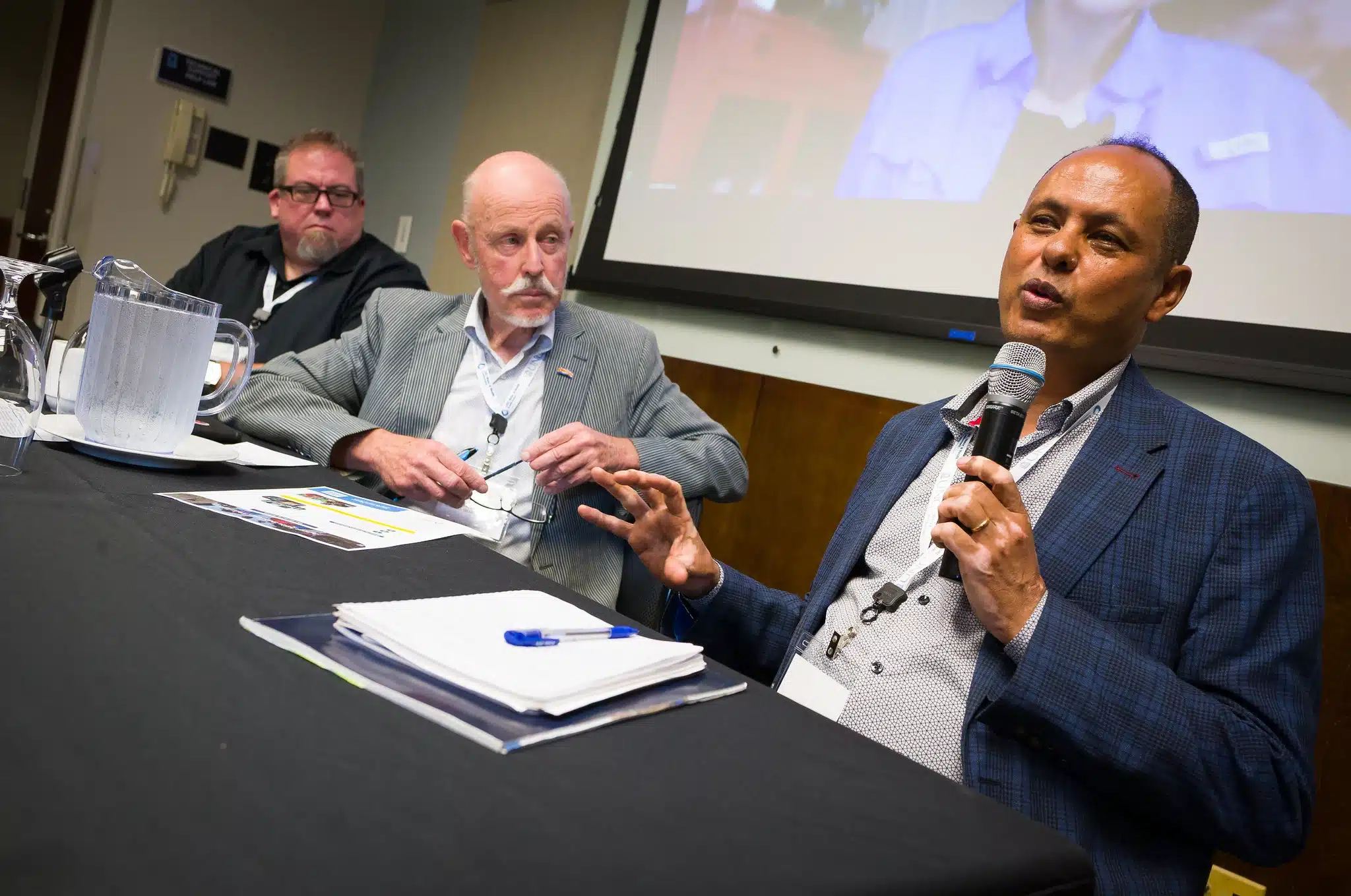 Panel discussion: a man on the right speaks into a handheld microphone while two other men listen, seated at a table with papers and a water pitcher in front of them.