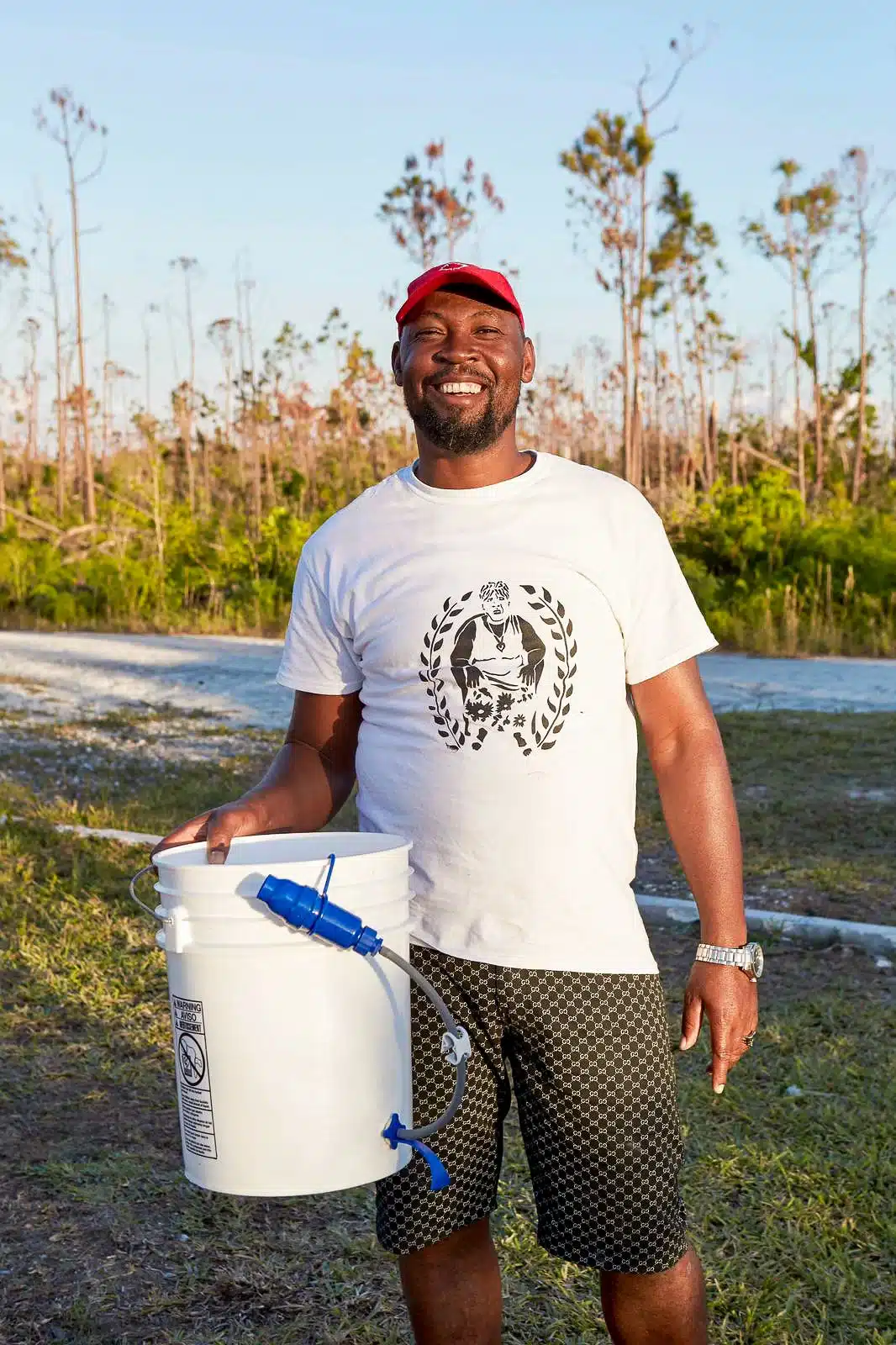 Smiling man in a white t-shirt and red cap holds a white bucket with a blue spigot outdoors in a sunny, sparse landscape.